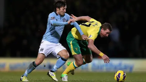 Getty Images Two male footballers battle for the ball with the player on the left wearing a light blue and white Manchester City kit and the other stumbling in a yellow and green Norwich City kit as he stumbled after being challenged. Spectators and advertising boards can be seen in soft focus behind them.