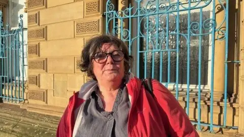 Gemma Dillon/BBC A lady wearing a red jacket and grey top is standing infront of some blue rails. 
She has brown short hair and glasses.