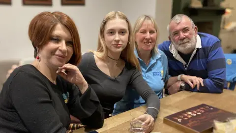 Three women and a man sitting in a row at a wooden table. The woman on the far left has chestnut-brown coloured hair, which is shaped in a short bob. She is wearing a black long-sleeved top and has her left hand under her chin with the elbow resting on the table. Next to her, on the right, is a teenager girl who has long blonde hair with clips on either side of her head. She is wearing a grey long-sleeved top, a black choker necklace and large silver hooped earrings in both ears, a silver bangle and has manicured nails. She is holding a glass, which is half-filled with clear liquid. On the right of the teenager is a mature woman with blonde hair and is wearing a blue polo t-shirt, her left arm is resting on the left of arm of a man sitting next to her on the right. He has grey hair and same coloured beard. He is wearing a blue and black striped top and is resting his right elbow and arm on the table.