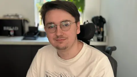 Jack is wearing glasses and a cream T-shirt. He is sitting in his wheelchair in the kitchen