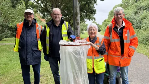 Elliot Deady/BBC Four people standing next to a bin which has been installed on a lamp-post. They are all smiling and wearing high visibility jackets.