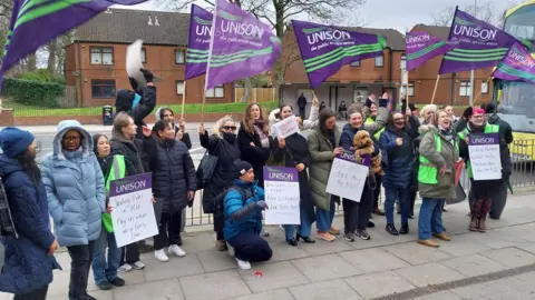Group of union members with UNISON flags stand on a curb.
