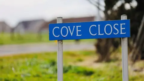 Blue street sign with words Cove Close in white capital letters, and housing in the background.