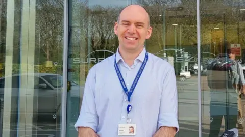 Andrew Cawthorne is standing and smiling in front of a glass entrance a building. He is wearing a blue shirt and lanyard and smiling at the camera.