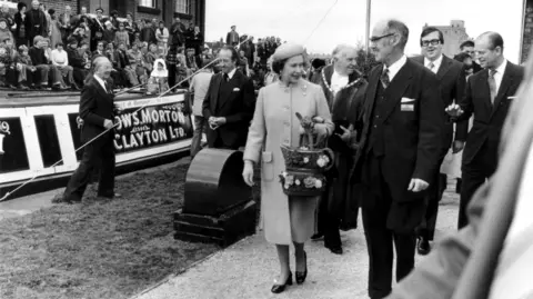 Canal & River Trust A black and white photograph of Queen Elizabeth II and Prince Philip walking past canal boats with a number of officials dressed in dark suits while a crown watches on.
