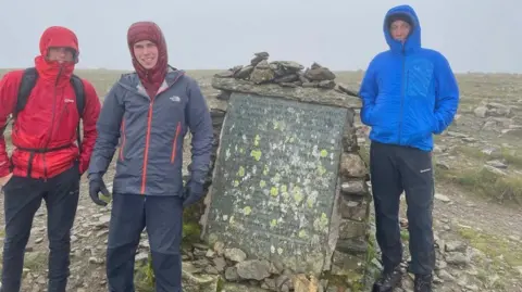 Matty Belcher | Brandan Smith Three young men in red, grey and blue winter jackets stand at the top of a cloud covered mountain top with their hoods pulled tightly around their faces. 