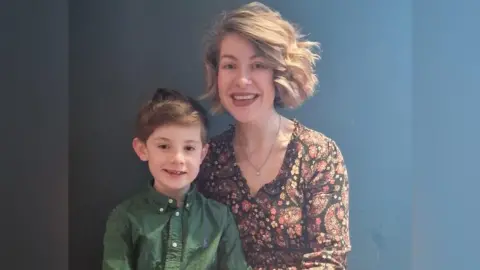 Bertie has brown hair and is wearing a green shirt. He is sitting with his mum Lauren, who has light brown short hair and is wearing a red and orange floral shirt.