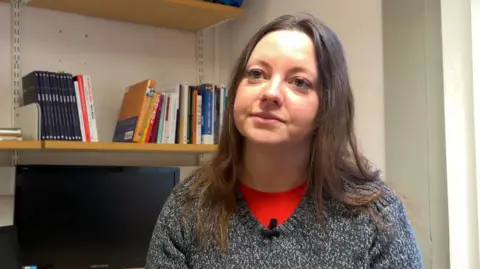 Ms Rutter is wearing a dark grey jumper and red t shirt. She has long, dark brown hair. She is pictured in her office at Durham University.