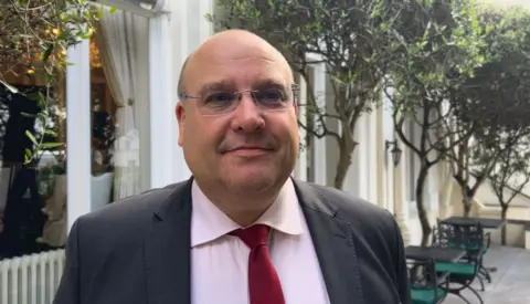 A bald man with rectangular glasses smiles at the camera in front of a hotel courtyard, with spindly green trees and trestle tables behind him. he is wearing a grey suit, with a white shirt and red tie. 