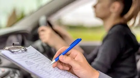 Getty Images A learner driver sat in a car, showing a driving instructor holding a pen towards a clipboard. 