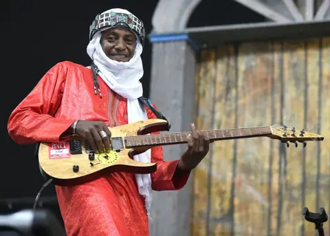 TIM MOSENFELDER / GETTY IMAGES A man dressed in red smiles as he plays the guitar, looking straight into the camera.