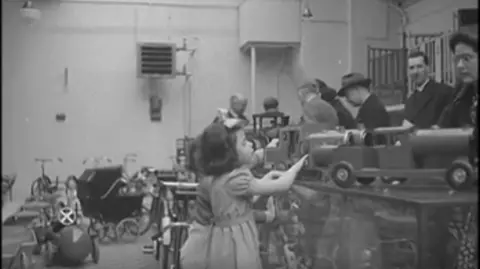 British Pathé A black and white archive image from 1945 of a young girl looking at toy trucks on a shop counter.