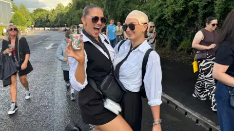 Two women. Both are wearing white shirts, black ties and sunglasses. The one on the right is wearing a bald cap and the one on the left is wearing a black waiscoat.