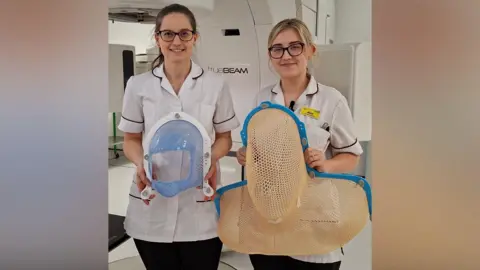 Two nurses are standing next to each other. On the left the woman has brown hair and glasses, and is wearing a white uniform. On the right the woman has blonde hair and glasses, with white uniform and a yellow badge. On the left the woman is holding a small mask that goes over someone's face - it has blue mesh that covers much of the patient's face but with a large hold for their nose and mouth. On the right, the mask is mesh and would cover the entirety of someone's head and shoulders