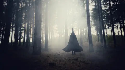 Getty Images A woman with black cloak and a hat, walking through a misty forest