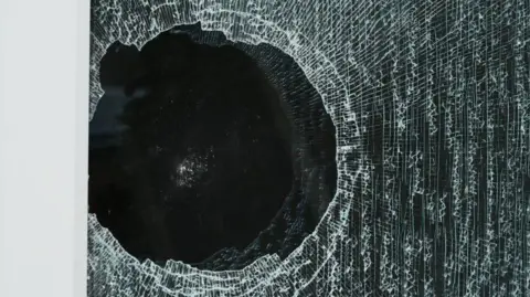 John Fairhall/BBC Broken window. It is a black, round hole with peripheral cracking around it.