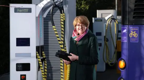 A woman with short brown hair stands in a bus depot in a black coat and pink scarf.