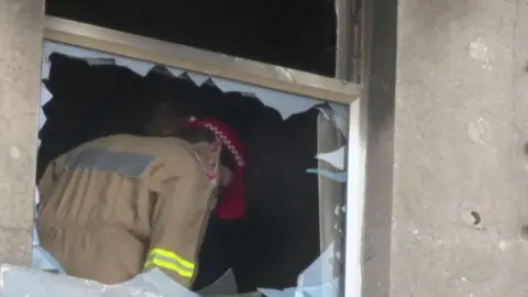 The picture shows a fire officer wearing a tan coloured uniform and red cap framed by the broken window of a flat. 