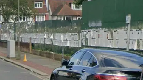 A Google Maps street view of the road in question with a series of posters pinned to a large green wall. 