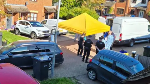 Submitted a large yellow tent and a white van, with police officers and people in full protective suits standing nearby. Several cars are parked around the scene in a quiet cul-de-sac.