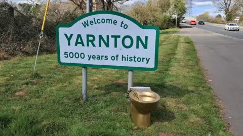 The gold toilet, next to a road and chained to a sign saying: Welcome to Yarnton, 5000 years of history
