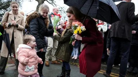 PA Media Kate crouching down smiling at a young child. Kate is wearing a long red dress and a burgundy jacket, holding several bouquets of flowers and holding a black umbrella. The young girl has a pacifier in her mouth and is wearing a pink coat with a polar bear on the pocket.