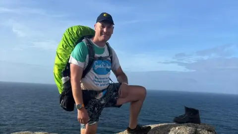Matthew Miles An image of a man wearing shorts, baseball cap and a Sue Ryder t-shirt and with a large green backpack on his back. He is standing with one leg on a rock. Behind him can be seen a calm blue sea and a blue horizon.