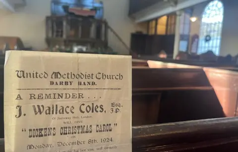 A sheet including the words Dickens Christmas Carol is upright among chapel pews. A window is on the right.