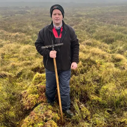 A man holding a rake stands in a field full of moss