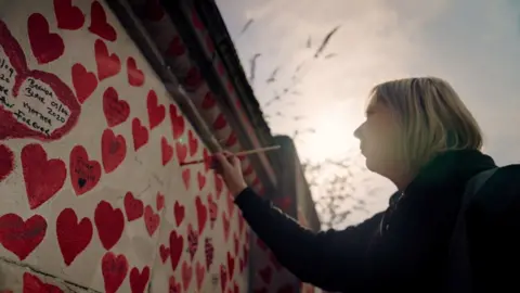 A woman, wearing a black coat and with short blonde hair, is painting a red heart on to the Covid memorial wall