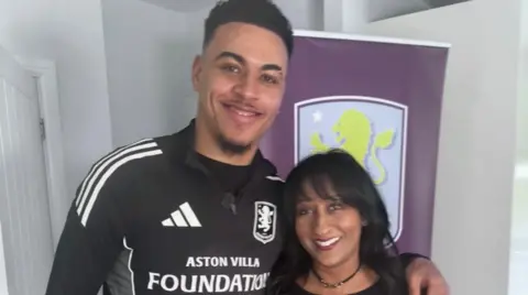 Morgan Rogers on the left has his left arm around Zena Weeks and they are both smiling at the camera. A large Aston Villa crest is behind them.