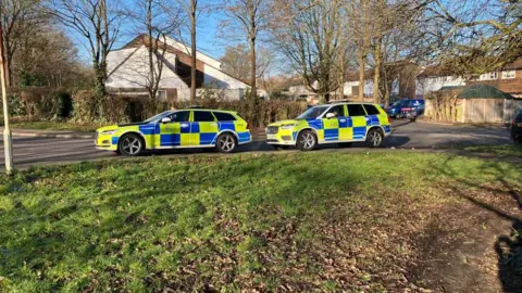 Sam Read/BBC Two marked police cars are parked on a residential road. There are houses in the background and a green area at the front