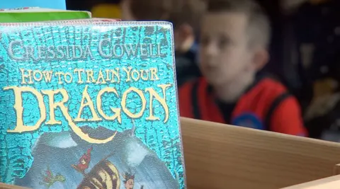 In the foreground there is a book called 'How to train your dragon'. In the background a child looks at someone out of focus.