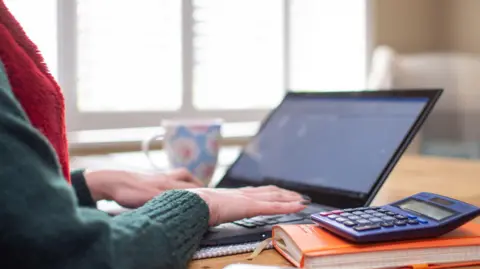 PA Media Woman tipos en computadora portátil en la mesa de la cocina, con una calculadora encima de un cuaderno