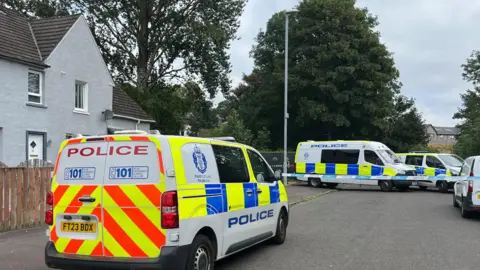 Police vans parked outside of a house in Glasgow. There is a police cordon and park with trees in the background.