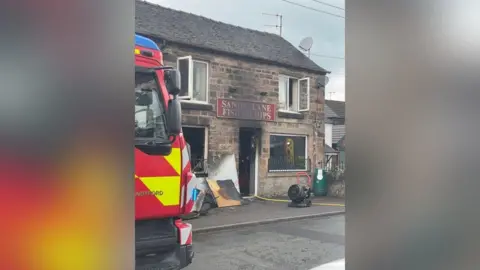 A brick building which has been badly burnt at the front. Charred wooden boards have been placed outside and a hose reel can be seen snaking inside through the open front door. A red sign above the front door says Sandy Lane Fish & Chips. The front of a fire engine is on the left.