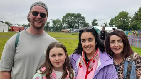 A family - a man, two women and a young girl - stand in a filed at the Stendhal festival.They are wearing clothes for an outdoor festival. They are smiling.