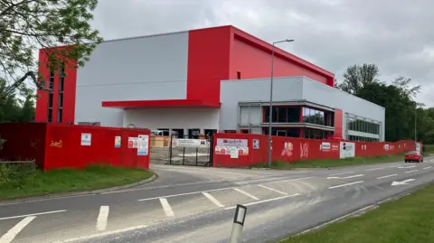 Amy Holmes/BBC A modern red-and-white building under construction sits behind a red site fence beside a road, with signage on the fencing and cars passing under a cloudy sky.