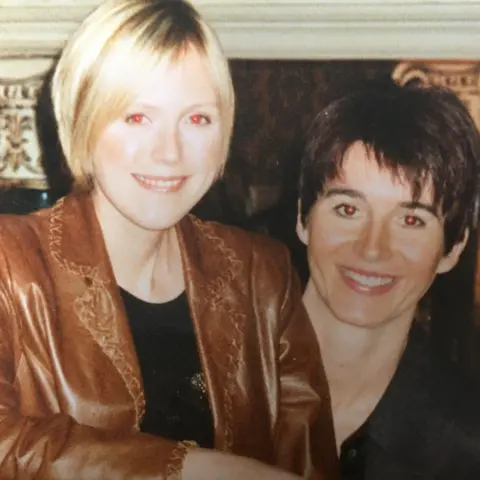 Angela MacVicar Family photo of smiling Johanna MacVicar, with short blonde hair and wearing a brown leather jacket, sitting next to Lindsay MacCallum with short dark hair and a black blouse