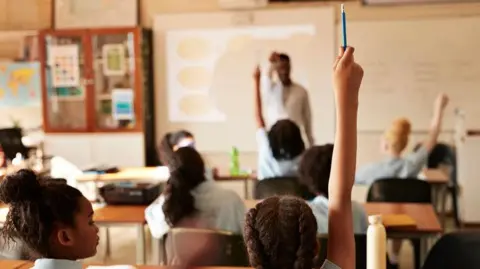 A primary school classroom with pupil sitting at desks. We can see a pupil in the foreground raising her hand with a blue pencil. We can only see the backs of the pupils heads. The teacher stands at the front of the class wearing a white shirt. The photo is partially blurred