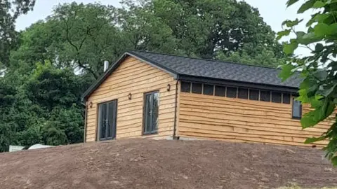 One of the homes built without permission by Michael Merrill. It is made from honey-coloured timber with black pitched roof. In the foreground is a mound of bare earth, and trees can be seen behind the building.