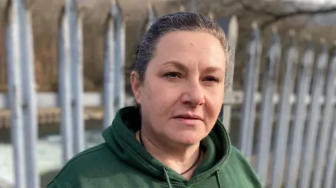 BBC Sharon Elward, local organiser for Pontypridd foodbank stares at the camera. She is standing in front of the river Taff in Pontypridd, wearing a green hoody. Metal railings can be seen behind her, and beyond them the river.  