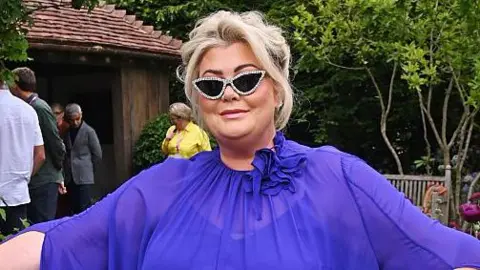 Getty Images Gemma Collins wearing a royal purple dress with her arms fanned out and the sleeves flowing in a garden at Chelsea Flower Show