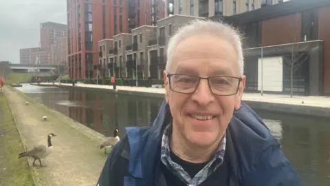 Al Franco - a trustee of the Manchester, Bolton and Bury Canal Society - stands by a canal and apartment buildings at Middlewood Locks in Salford. Geese can be seen walking behind him.