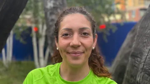 A woman - Renata Wais - smiling directly at the camera. She is standing in front of a wooden sculpture and is wearing a green running T-shirt.