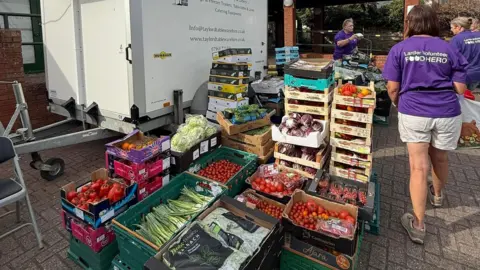 Towcester Community Larder A large selection of boxes of fresh food, all piled up, next to three people who are wearing purple T-shirts with the words Larder Volunteer Food Hero in white writing on the back. There is a white trailer unit to the left. 
