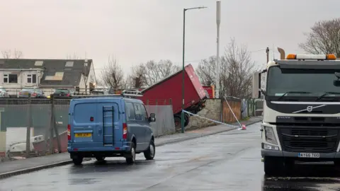 The rear end of a red lorry pointing upwards after crashing into a fence