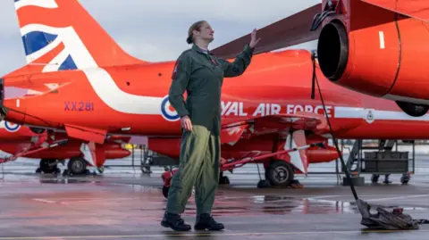 MOD / PA A female pilot, dressed in a green flight suit, inspects a Red Arrow jet on the tarmac.