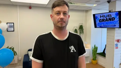 John Penn wears a black and white t-shirt and stands in front of a TV screen which says "Hub grand opening". A sofa is in the background.