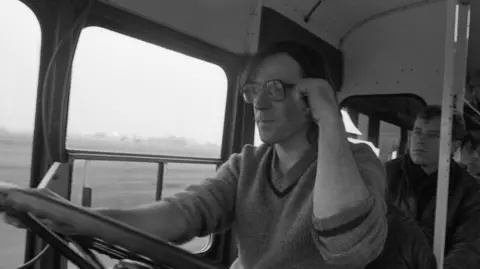 Family photo A black and white photo of a man with long dark hair and glasses holding a large steering wheel with the interior of a bus behind him